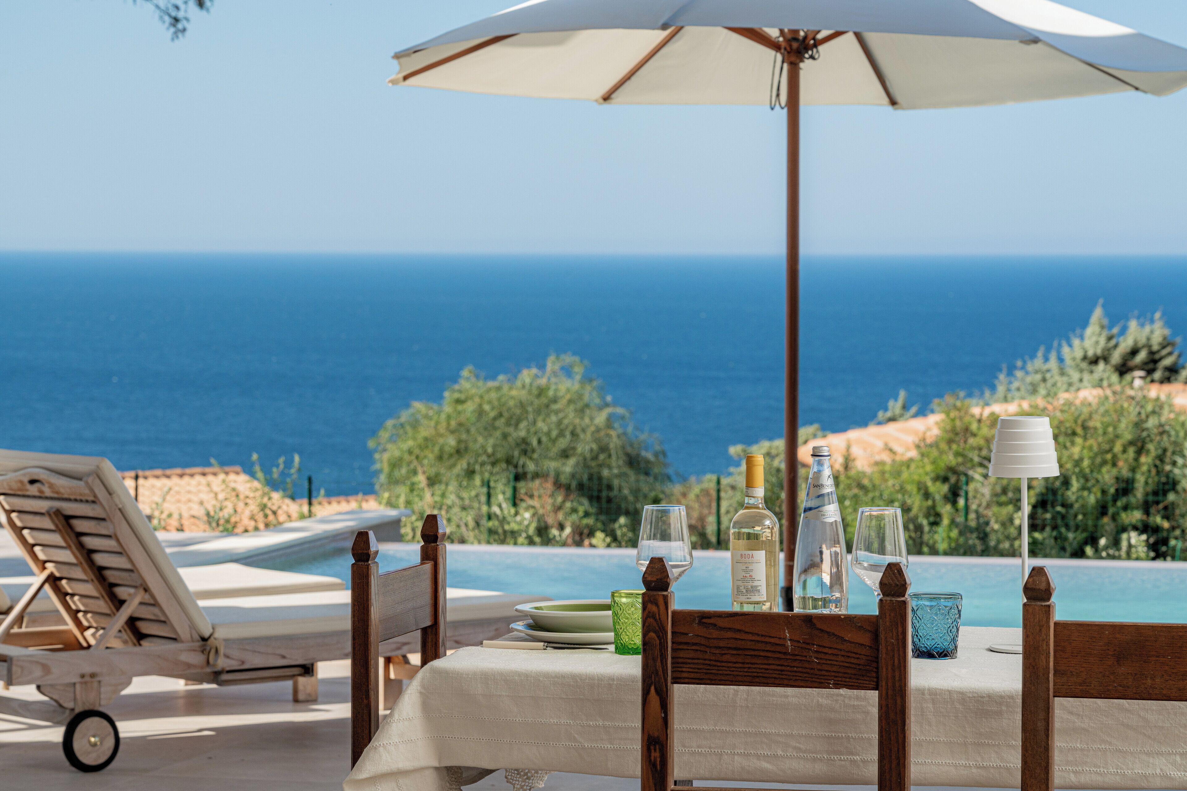 A dining table with a white tablecloth, a bottle of wine, and glasses, set against a backdrop of a pool and ocean view.