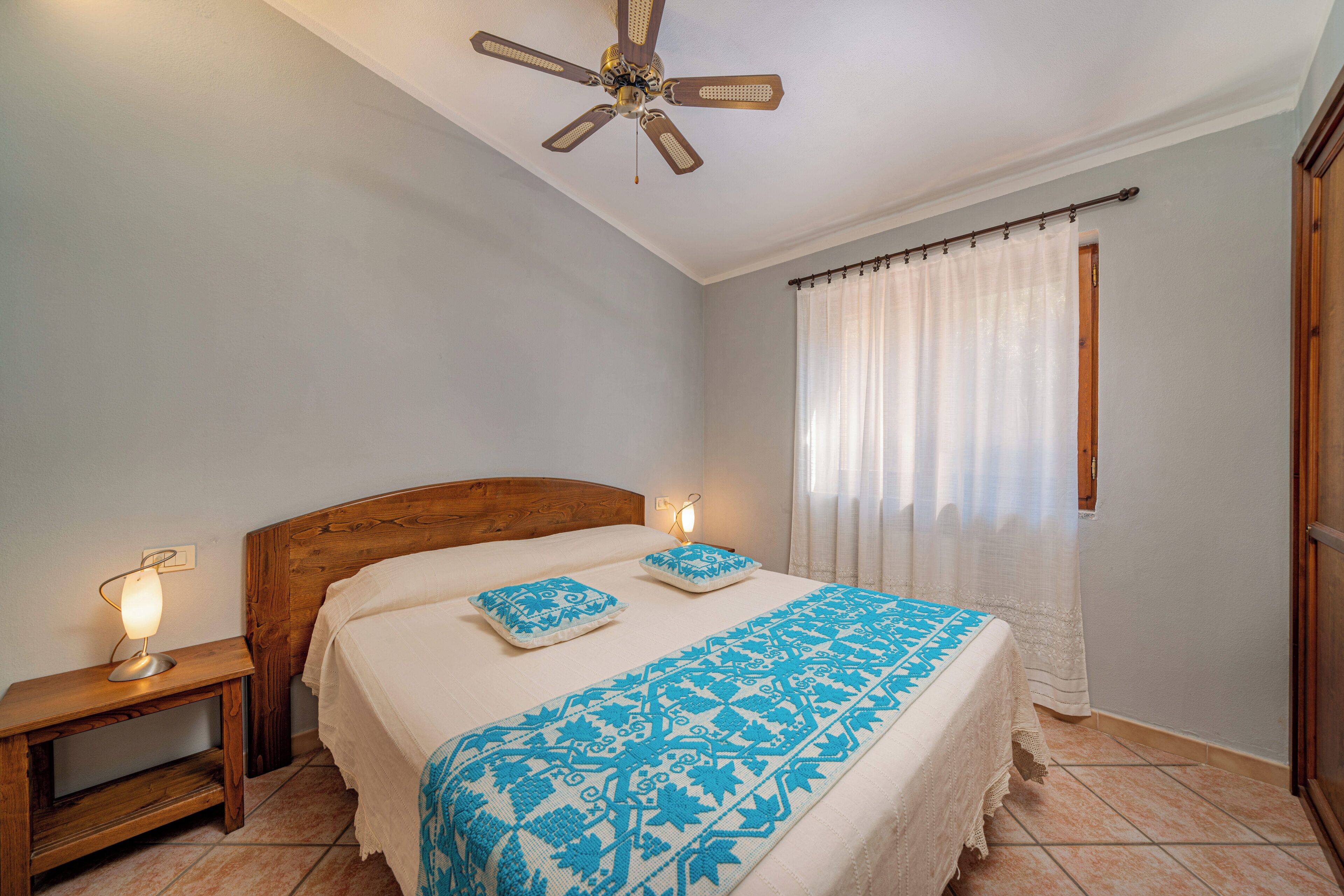 A bedroom with a wooden headboard, a bed with a blue patterned bedspread, a nightstand with a lamp, and a ceiling fan.
