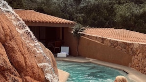 A pool with a waterfall feature, surrounded by a rustic stone wall and a small building with a tiled roof.