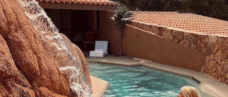 A pool with a waterfall feature, surrounded by a rustic stone wall and a small building with a tiled roof.