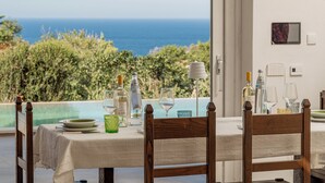 A dining table set for two with a view of the ocean, a white tablecloth, and various glassware and bottles.