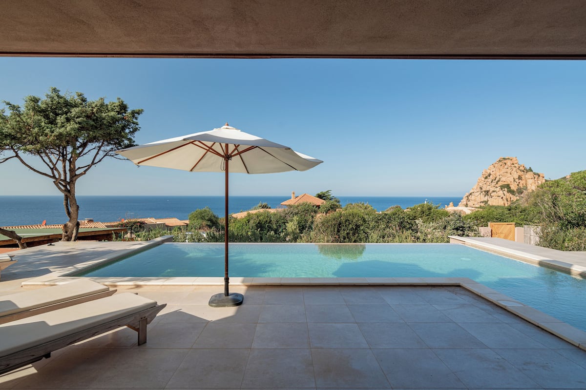 A pool area with a view of the sea, a tree, and a white umbrella.