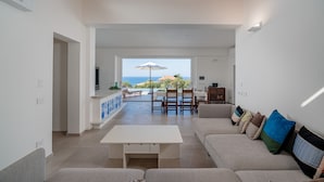 A spacious living area with a view of the sea, a dining area with a table and chairs, and a kitchen visible in the background.