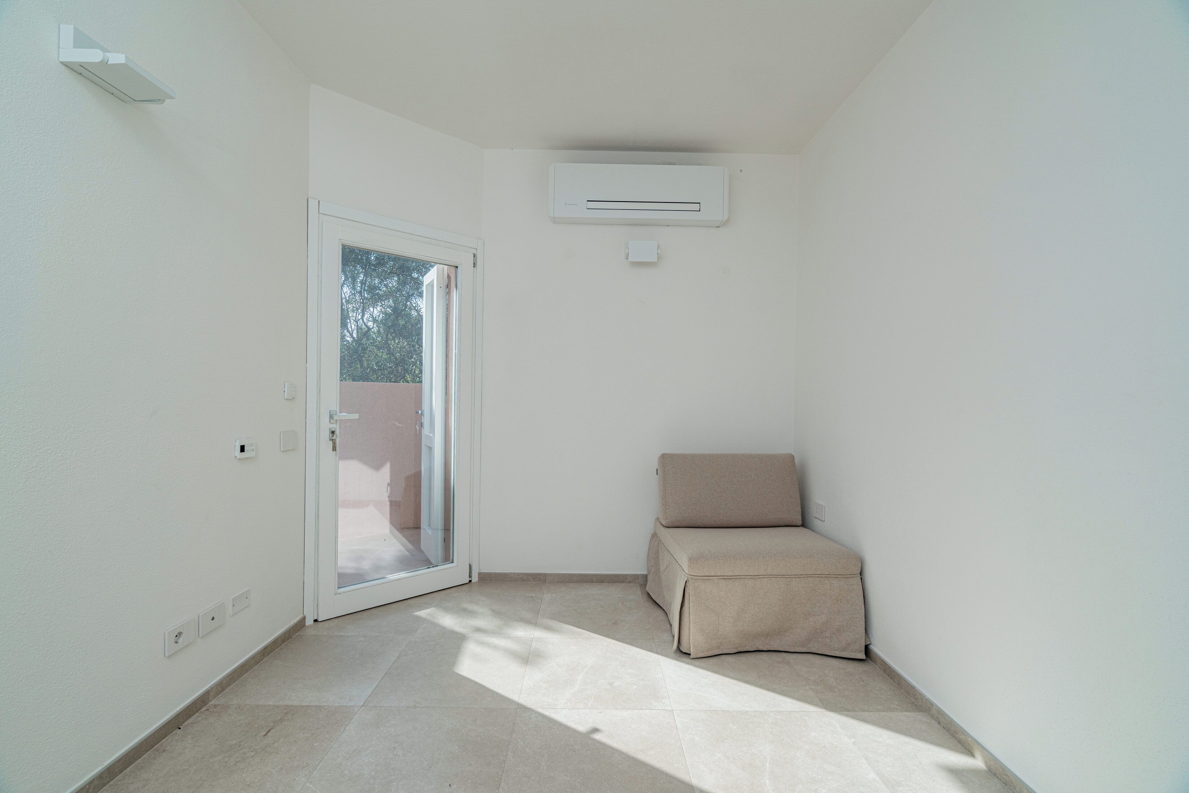 Una habitación pequeña y bien iluminada con un ventanal corredero que da a un espacio al aire libre, un sofá color beige y un equipo de aire acondicionado instalado en la pared.