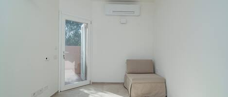 Una habitación pequeña y bien iluminada con un ventanal corredero que da a un espacio al aire libre, un sofá color beige y un equipo de aire acondicionado instalado en la pared.