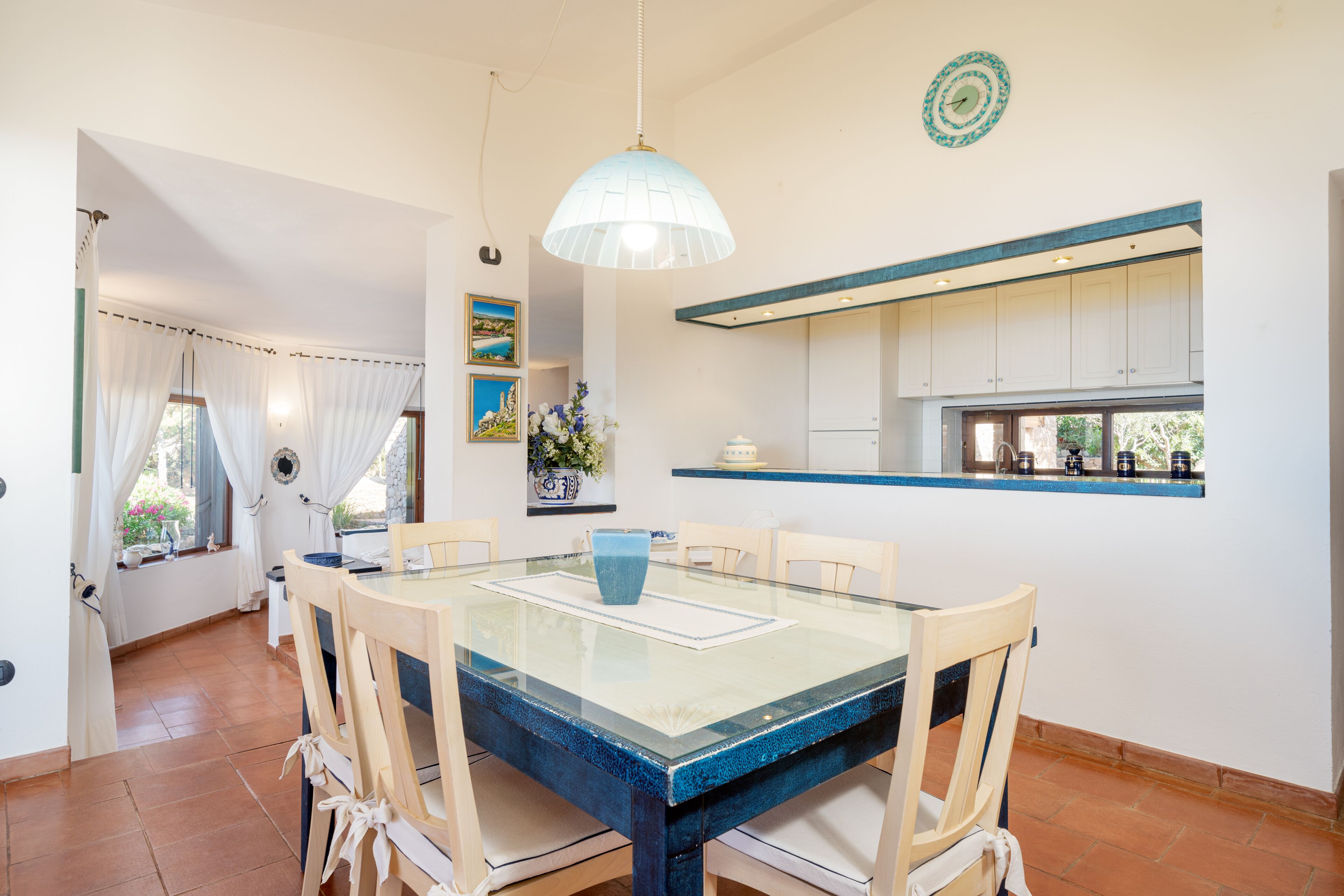 A dining area with a glass-top table, chairs, a hanging light fixture, and a kitchen with white cabinets and a window.
