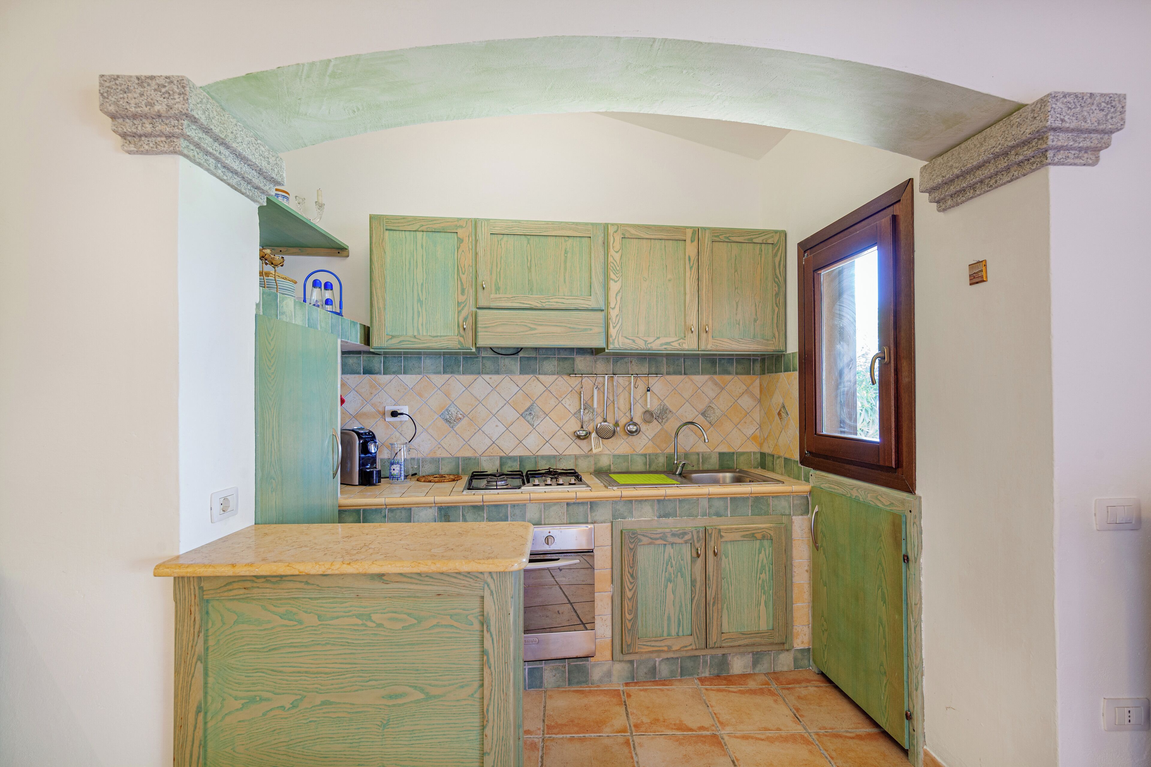 Une cuisine compacte avec des meubles de rangement en bois vert, un four encastré et une fenêtre au-dessus de l’évier.