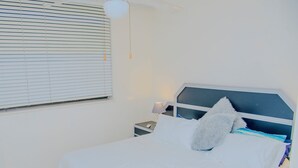 Een net opgemaakt bed met een zacht kussen, een nachtkastje met een lamp, en een raam met luxaflex.
