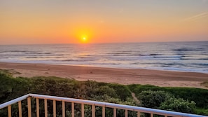 A beach with the sun setting over the ocean, a wooden railing in the foreground, and greenery in the background.