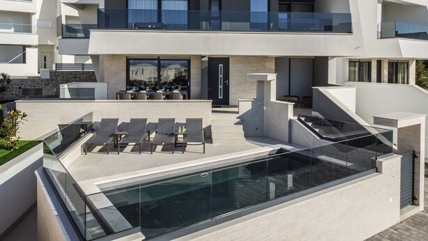 A modern two-story house with a rooftop pool, outdoor seating, and a stone wall.