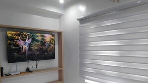 A modern living room with a mounted TV on a wooden shelf, a white fluffy rug, and horizontal blinds on the window. - Oceanview Studio Bacolod City (Bacolod)