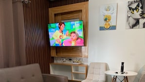 A modern living room with a beige sofa, a white armchair, a small round table, and a flat-screen TV mounted on a wooden panel. - Oceanview Studio Bacolod City (Bacolod)