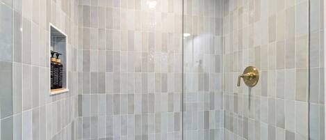 A shower with a glass door, a gold showerhead, and a shelf with toiletries.