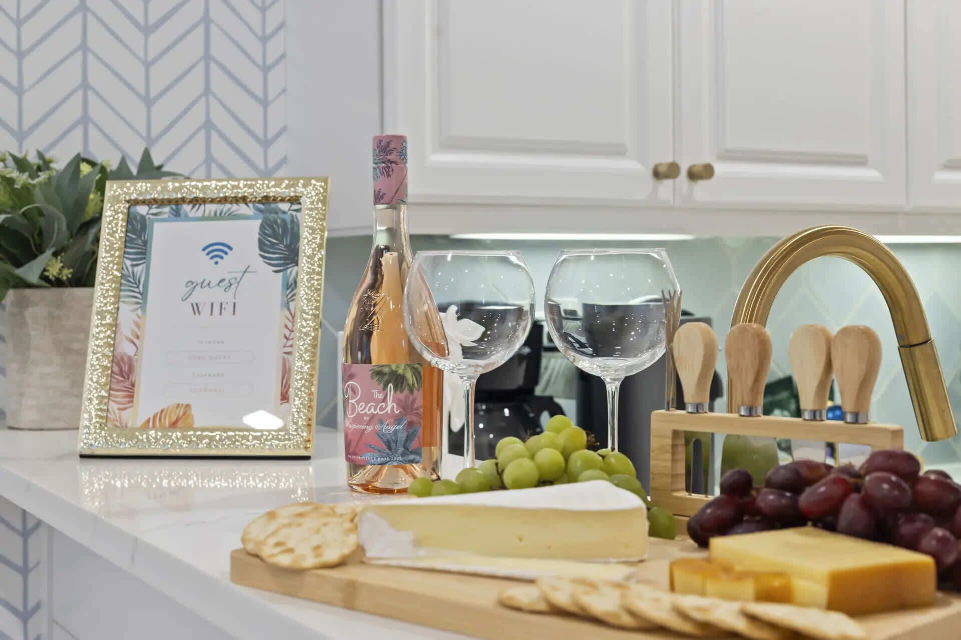 A kitchen counter with a framed certificate, a bottle of wine, two wine glasses, a cheese board with various cheeses and grapes, crackers, and a knife block.