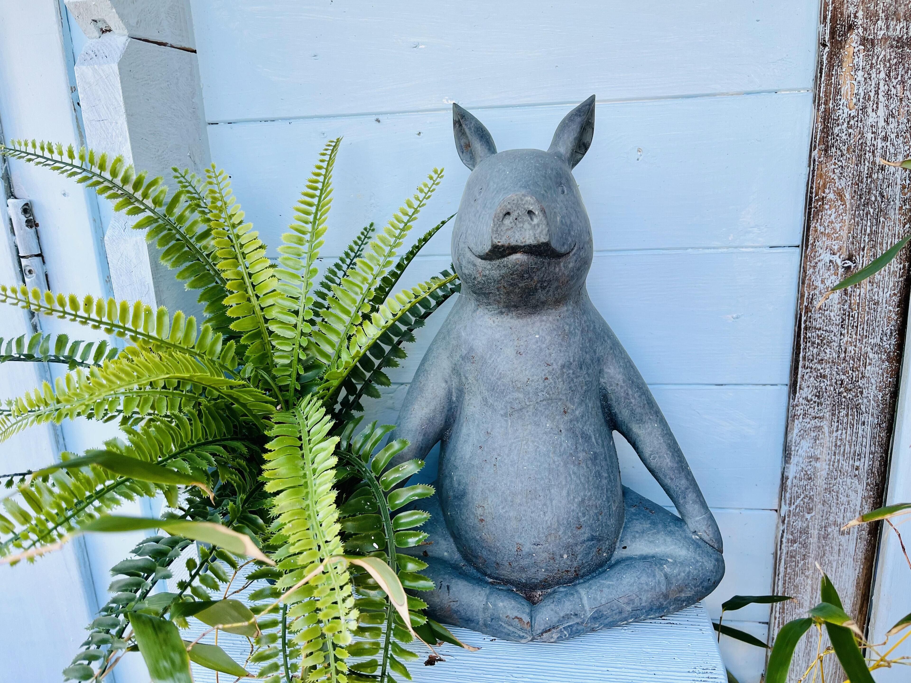 Une sculpture représentant une figure assise, dotée d’un corps arrondi et d’un dos plat, placée contre un mur blanc présentant des signes d’usure et une texture rustique.