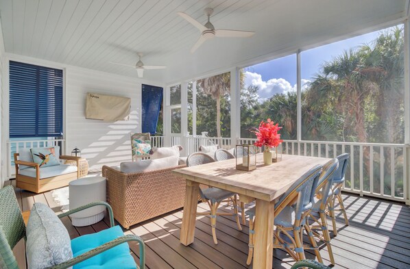A covered outdoor deck with wicker furniture, a table with a vase of flowers, and a view of tropical vegetation. - Award-Winning Resort Amenities | Yacht Harbor Court 4 (Isle of Palms)