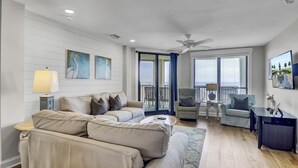 A modern living room with a beige sectional sofa, a small round coffee table, and a blue accent rug. There are two armchairs, a side table with a lamp, and a painting on the wall.