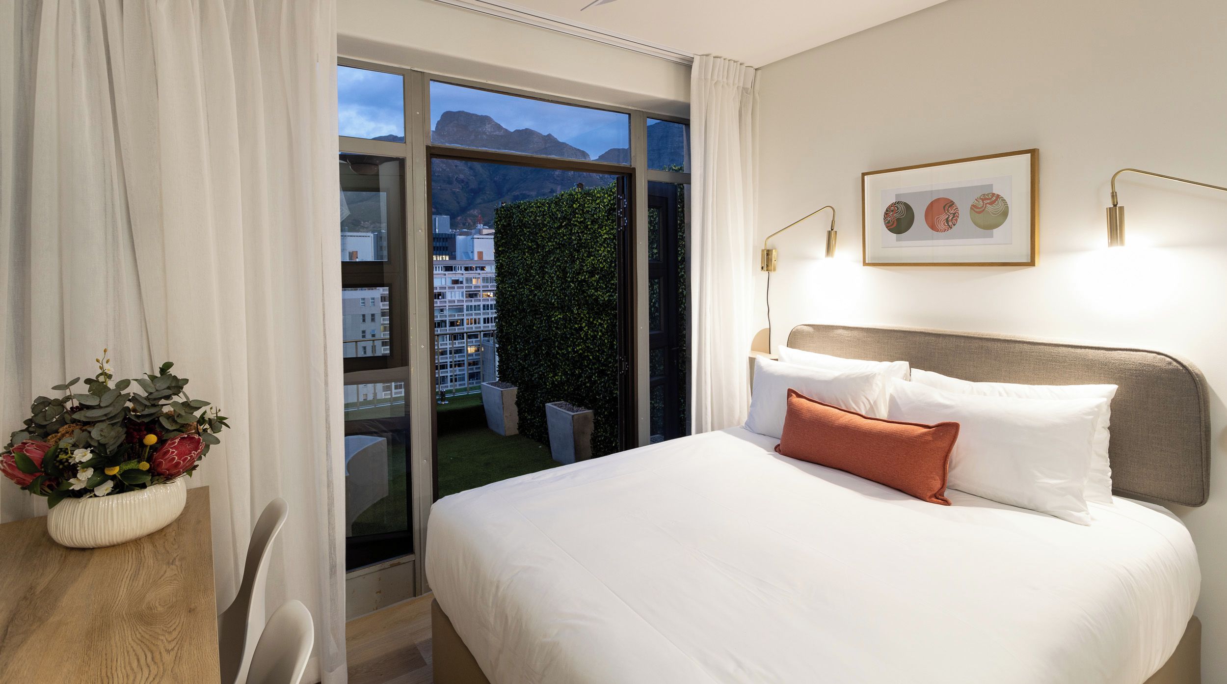 Ein Hotelzimmer mit Bett, Schreibtisch, Stuhl, Fenster mit Blick auf Gebäude und Berge sowie einem gerahmten Bild an der Wand.