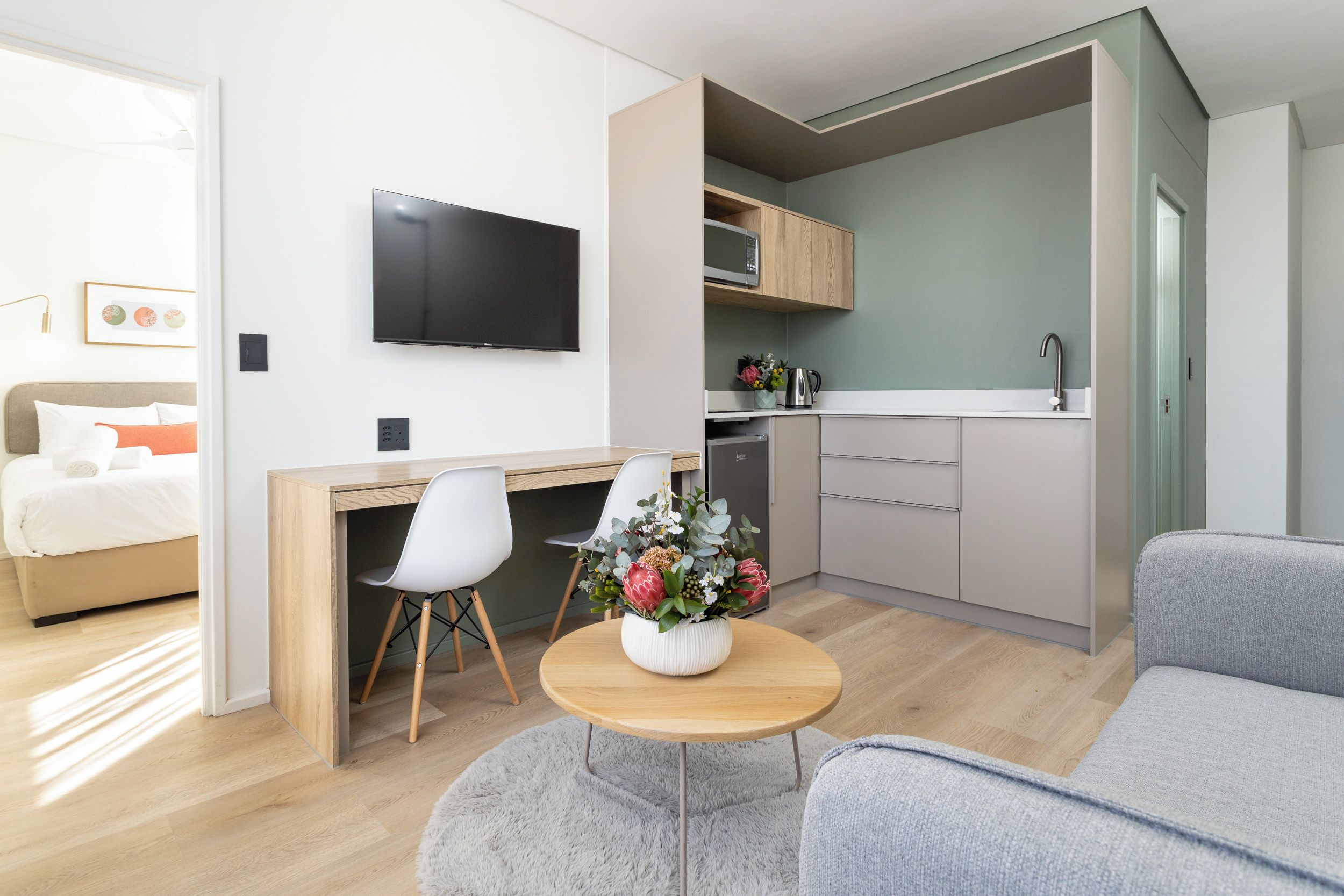 Un espacio habitable moderno y funcional con cama, escritorio, televisor de pantalla plana, kitchenette y sofá gris.