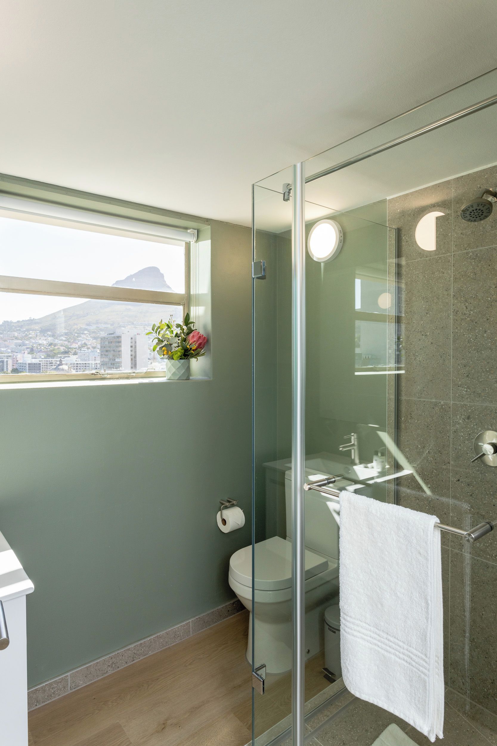 Un baño moderno con ducha de vidrio, inodoro y lavamanos. Se ve una ventana con vistas a un paisaje urbano.