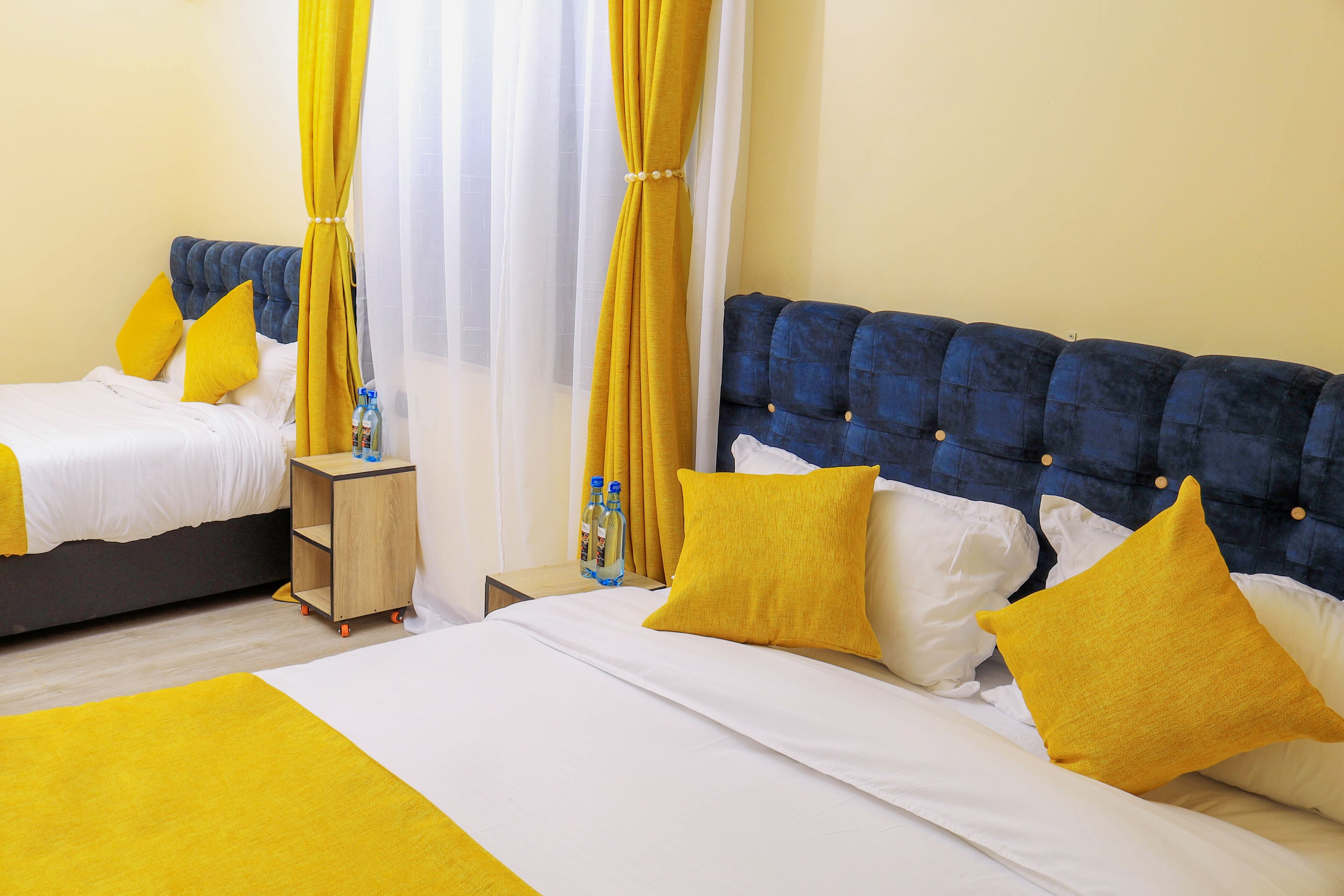 A hotel room with two beds, each with yellow and white bedding, a wooden side table, and a blue headboard.