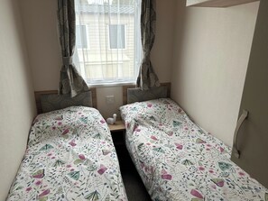 Ein kleines Zimmer mit zwei Einzelbetten, einem fensterbelaubten Fenster und einem an der Wand befestigten Lichtschalter.
