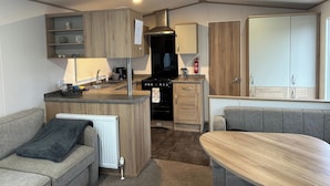 A compact kitchen with wooden cabinets, a stove, and a dining area with a table and chairs. - Felixstowe Chimo Caravan 8 Sleeps 5 min to Beach (Felixstowe)