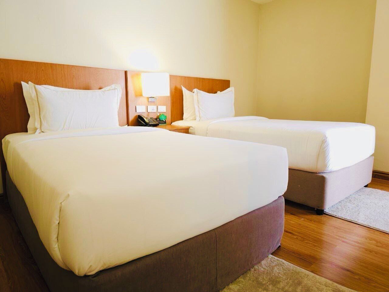 A hotel room with two beds, wooden headboards, a nightstand with a lamp, and a small table with a phone and a green object.