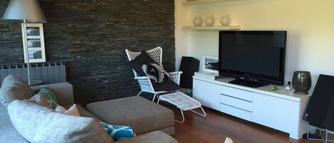 A modern living room with a grey sofa, a white armchair, a flat-screen TV, and a bookshelf with decorative items.