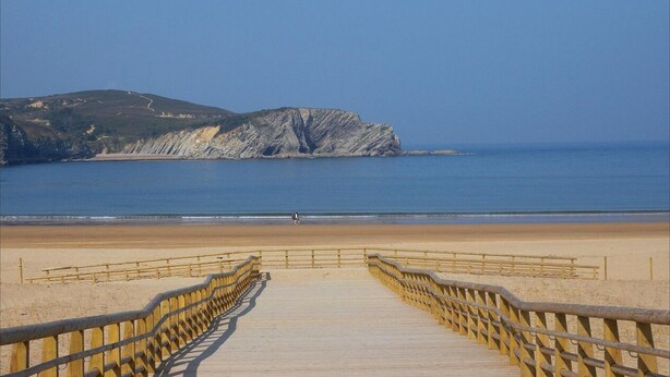 Une promenade en bois mène à une plage, avec une falaise rocheuse en arrière-plan.