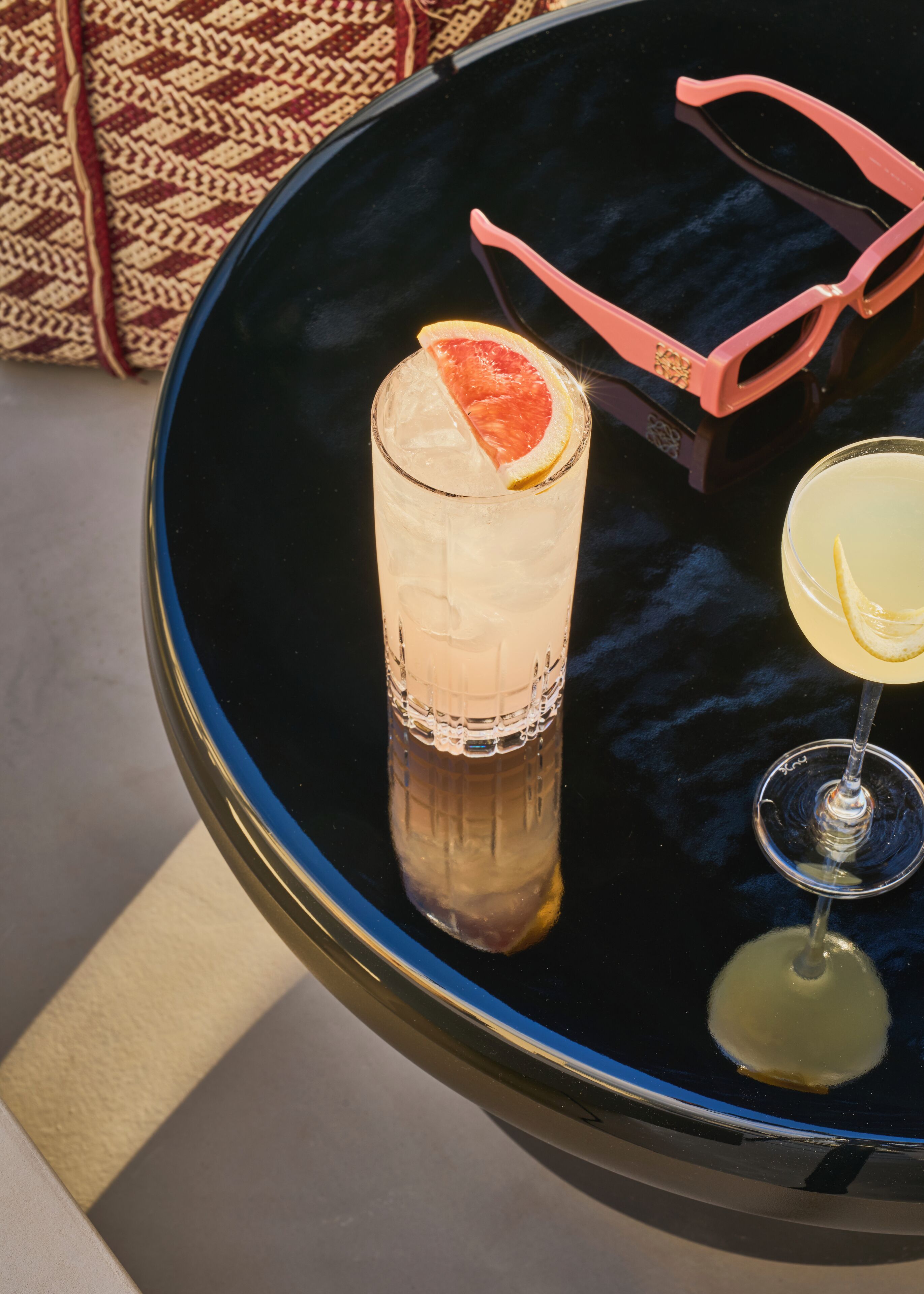 Uma mesa com um copo alto contendo uma bebida em camadas, uma taça de cocktail com uma fatia de limão e um par de óculos de sol rosa.