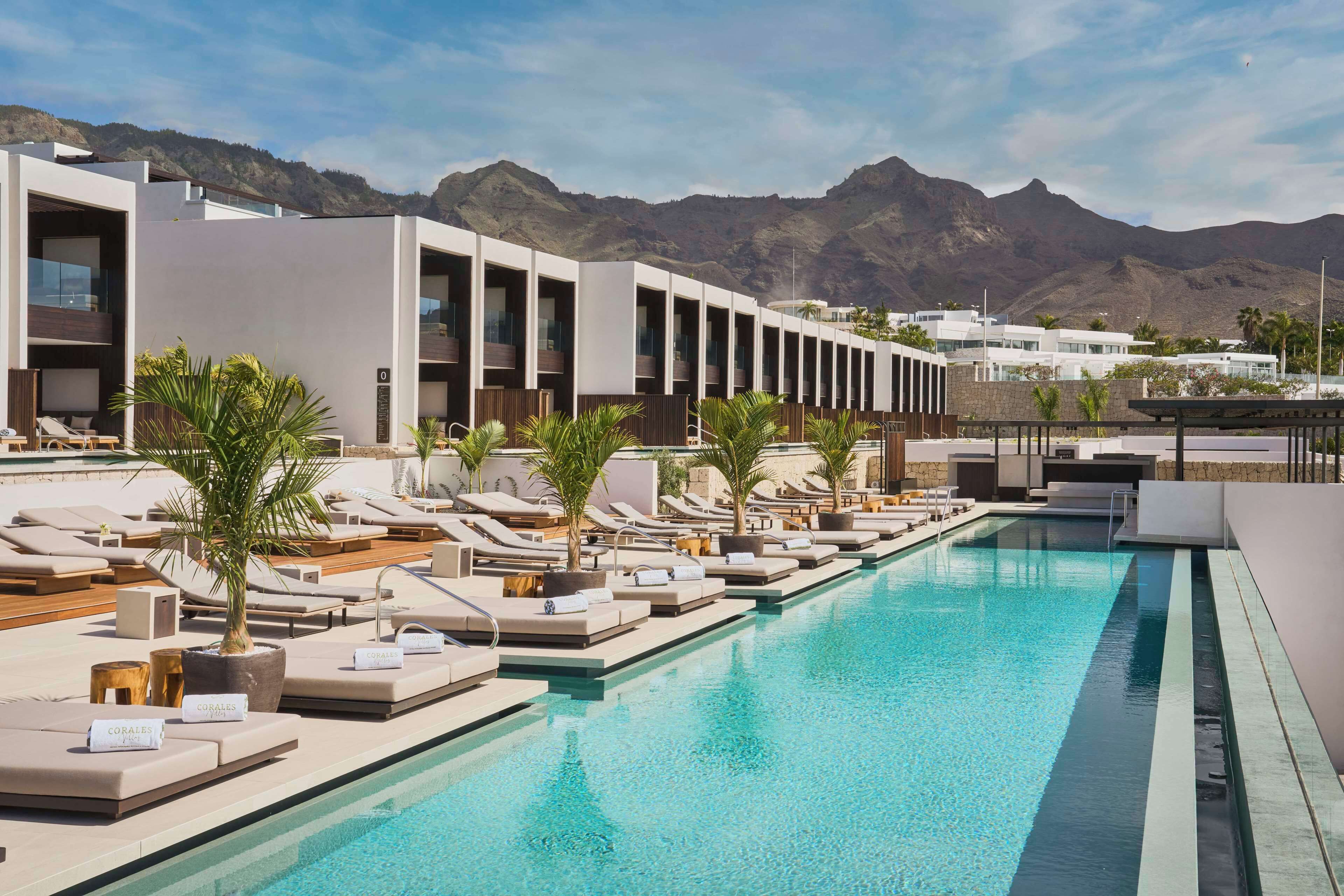 Zona de piscina moderna de hotel con sillones de descanso y palmeras, con montañas al fondo.
