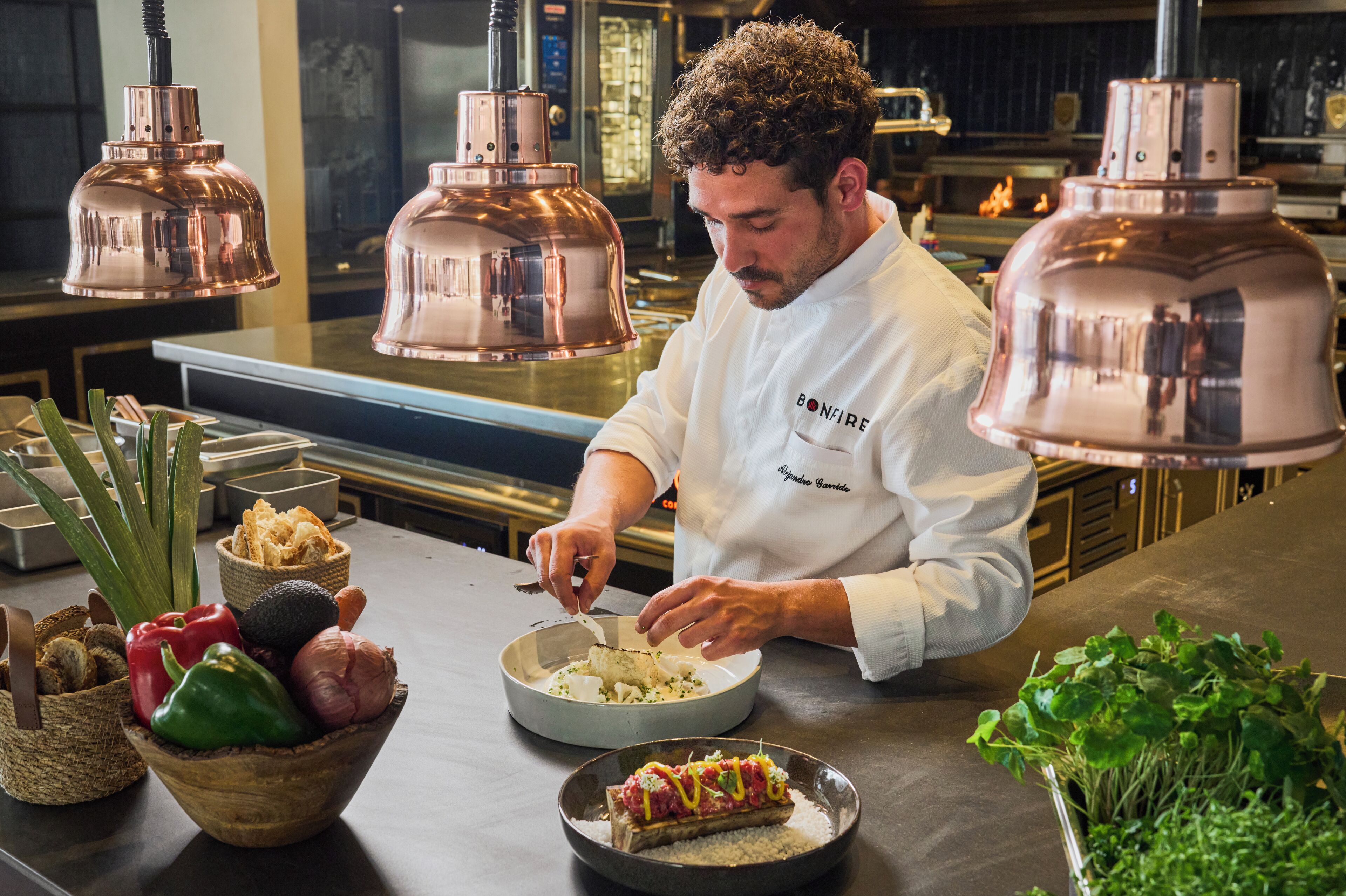 Un chef prepara comida en una cocina con lámparas de cobre e ingredientes diversos sobre la encimera.