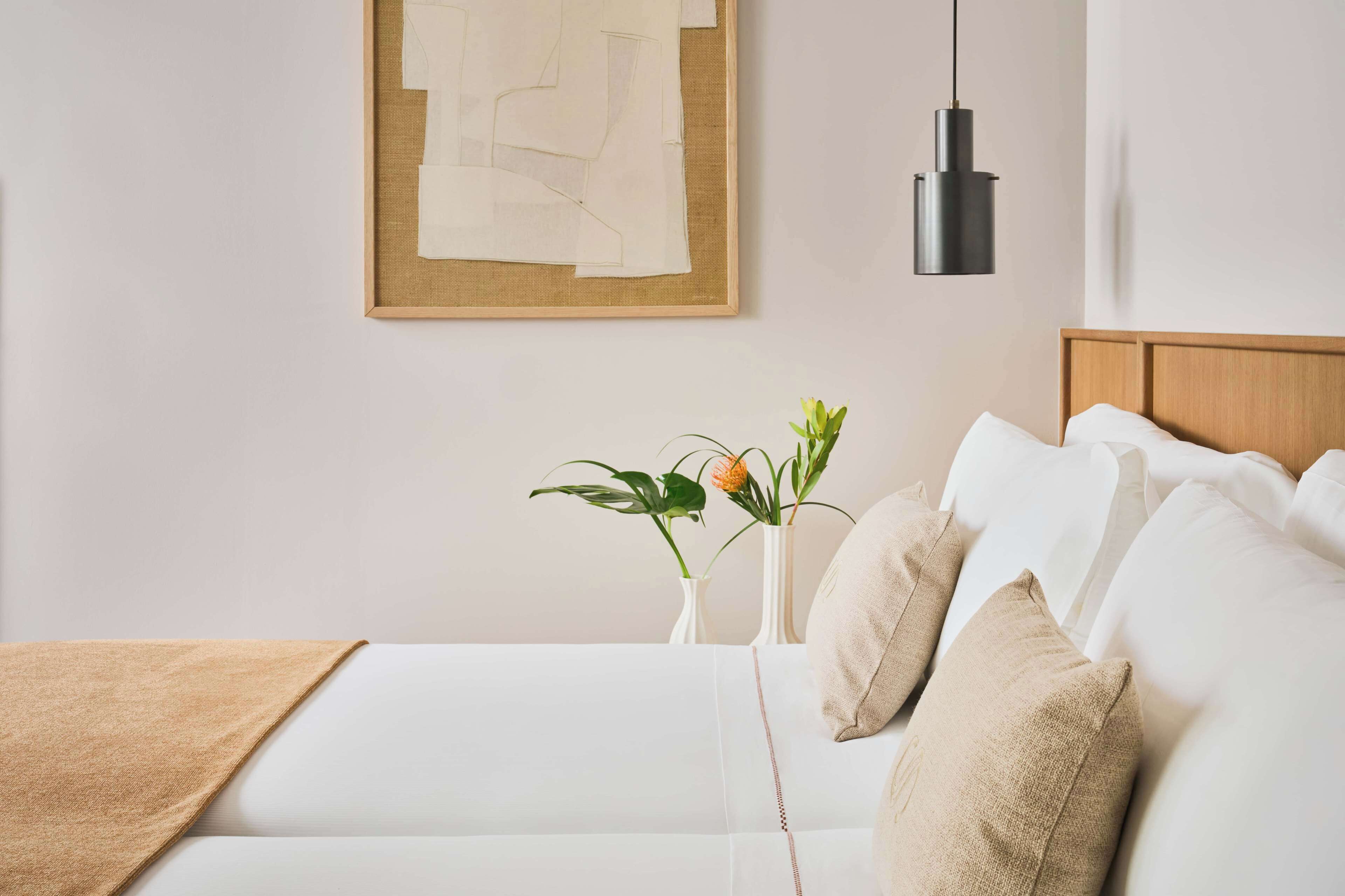A neatly made bed with white and beige bedding, a framed abstract artwork on the wall, and a hanging pendant light.