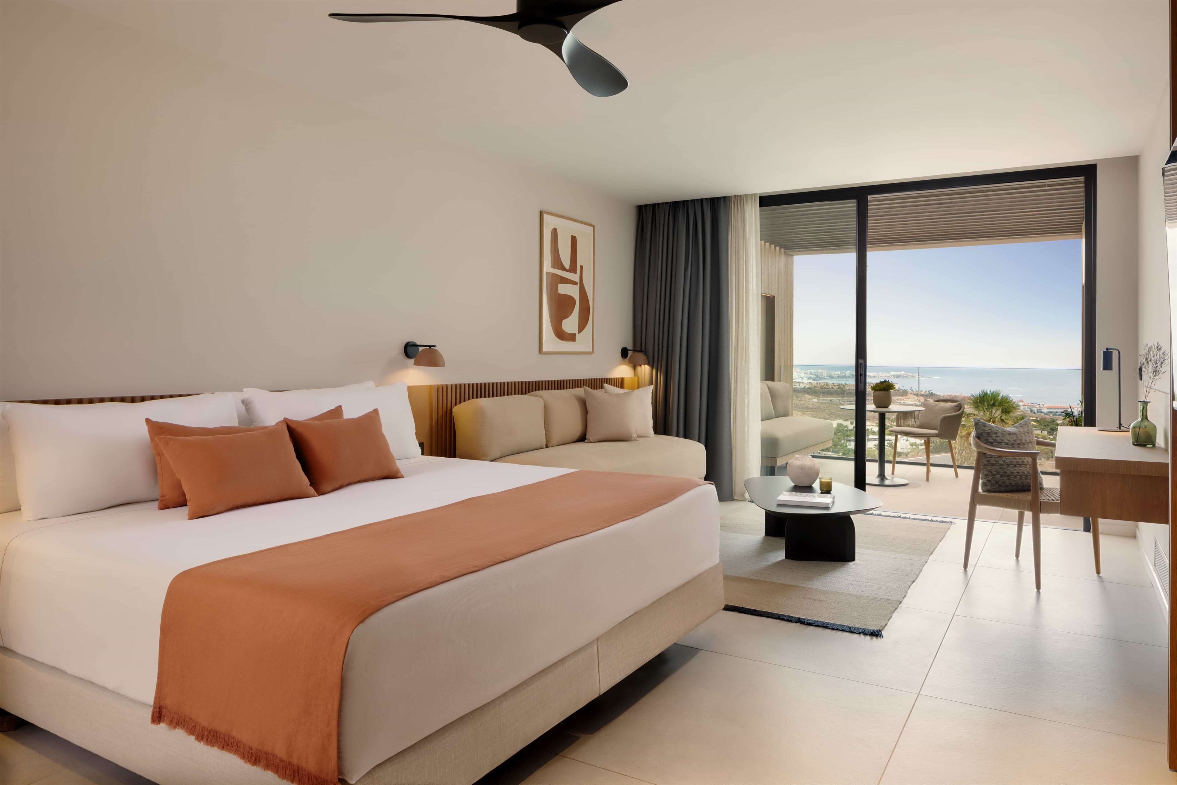 A modern hotel room with a large bed, a sofa, a dining area, and a balcony with a view.