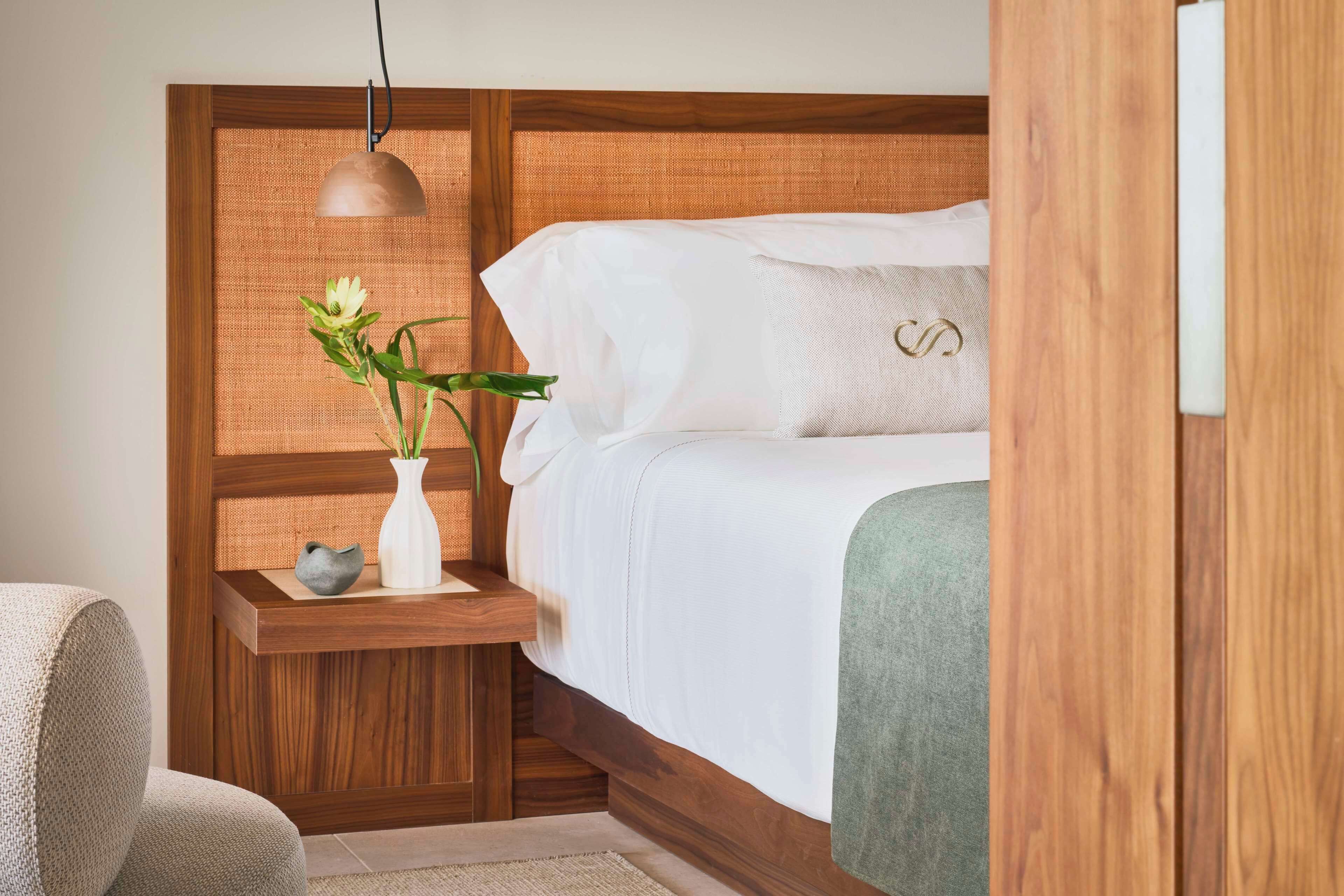 A neatly arranged bedroom with a wooden headboard, a bedside table with a vase and a bowl, and a hanging lamp.