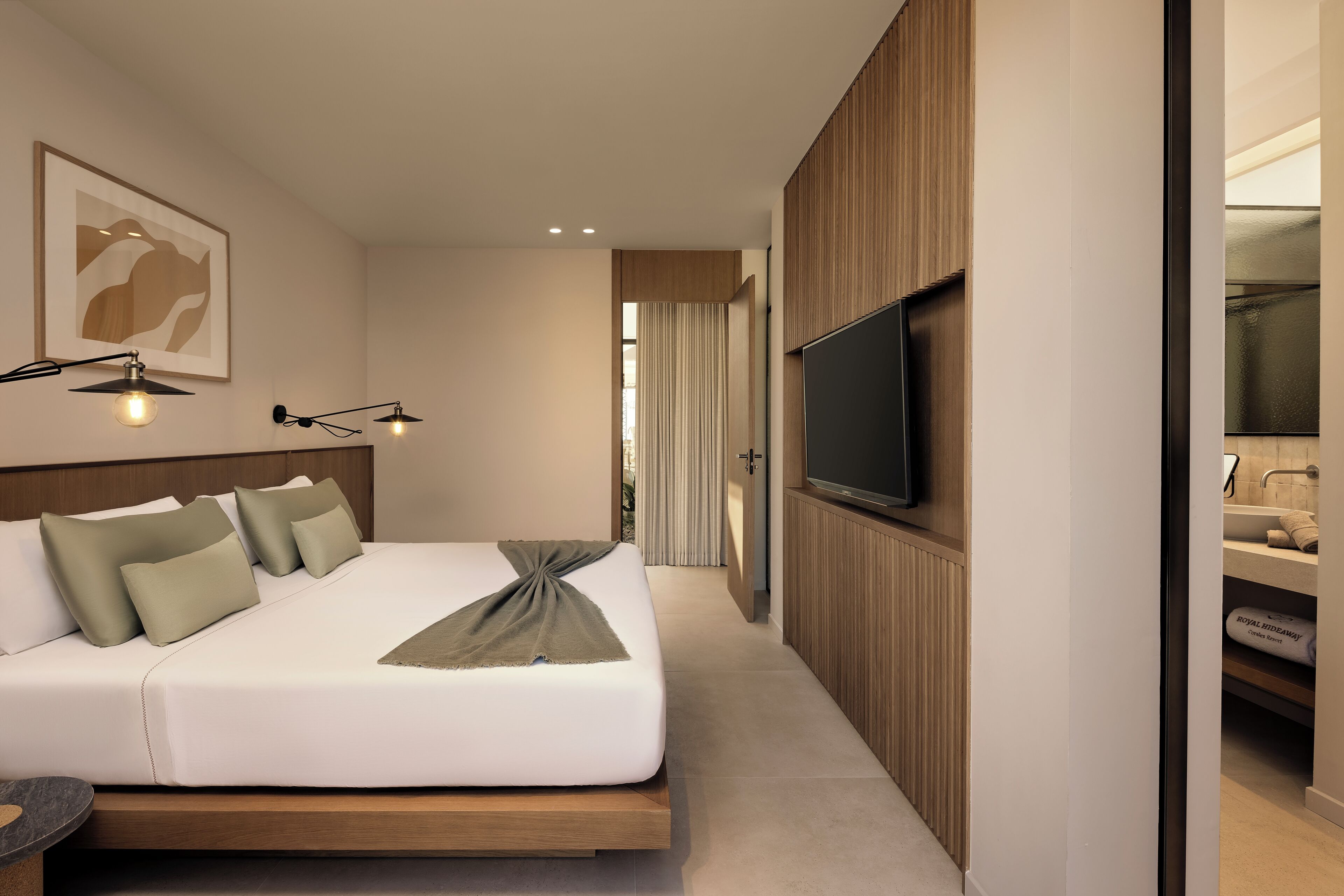 Una habitación de hotel moderna con una cama grande, un televisor en la pared y un baño con lavamanos y espejo.