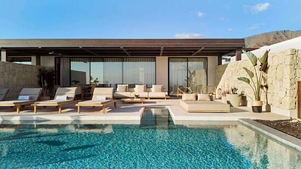 A modern outdoor pool area with lounge chairs and a sofa, surrounded by large windows and a stone wall.