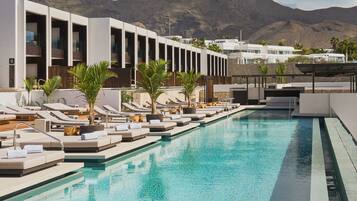 Una piscina con tumbonas y un edificio moderno con balcones, con montañas al fondo.