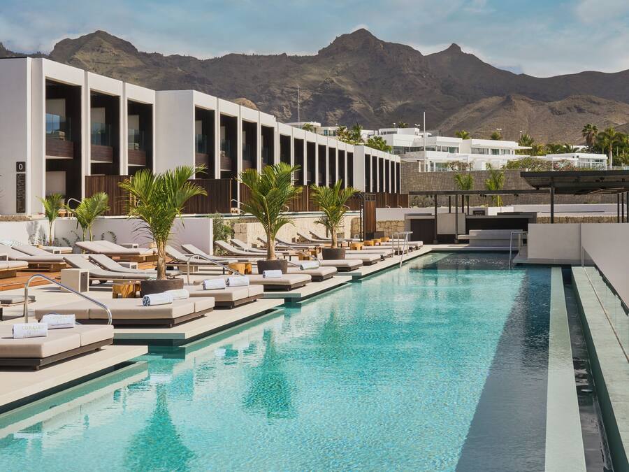 Una piscina con tumbonas y un edificio moderno con balcones, con montañas al fondo.