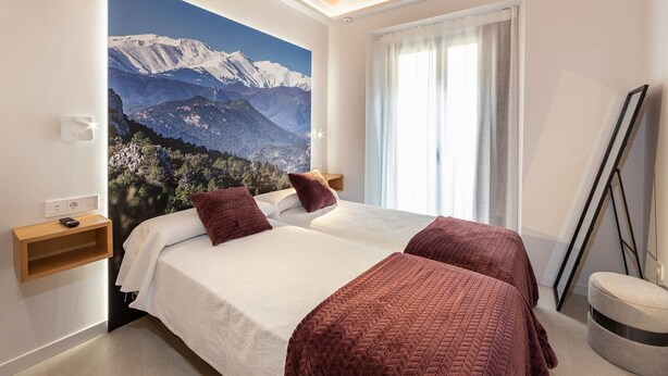 Une chambre dotée d’un grand lit, de deux tables de chevet, d’une fenêtre avec des rideaux et d’une fresque représentant un paysage de montagne.
