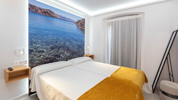 Ein Hotelzimmer mit Bett, Nachttisch, Fernbedienung und einem großen Fenster mit Blick auf ein Gewässer und eine felsige Landschaft.