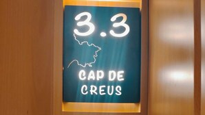 Ein Leuchtreklame mit der Zahl 3,3 und dem Text "CAP DE CREUS" an einer Holztür.