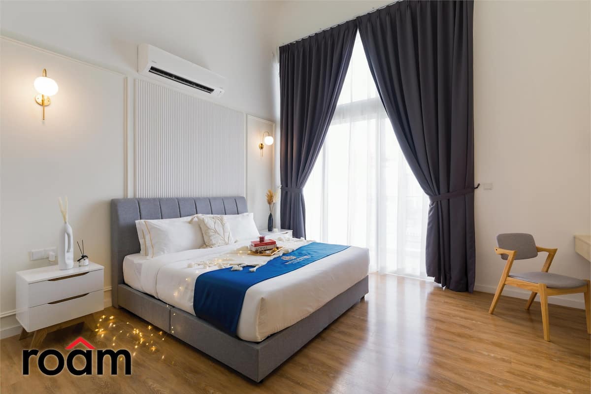 A modern bedroom with a grey upholstered bed, white bedding, a blue throw, a bedside table with a lamp, and a chair.