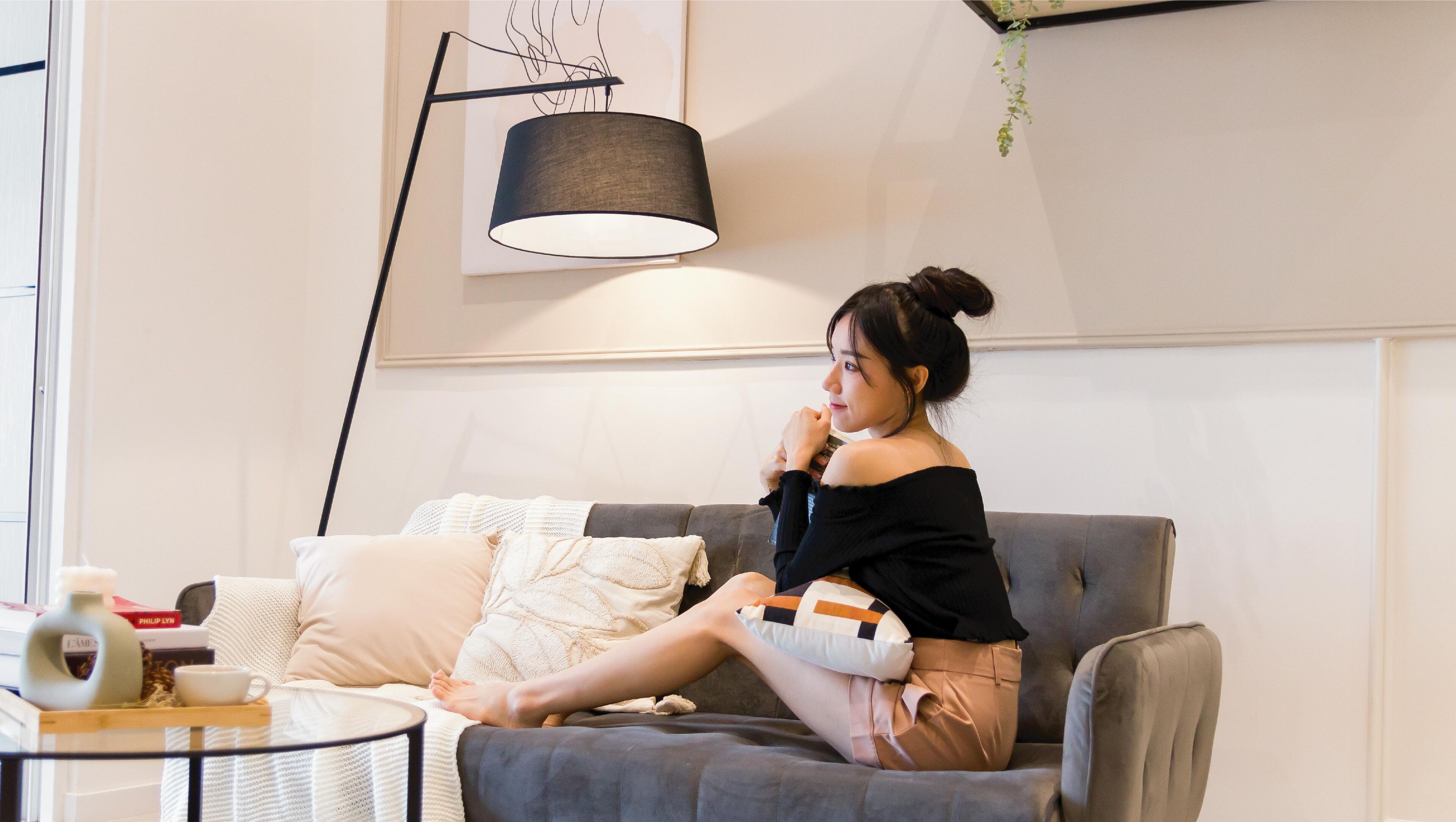A person sitting on a grey sofa with a white cushion, a black lamp, and a small table with a cup and a book.