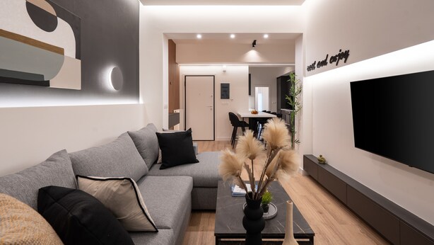A modern living room with a grey sectional sofa, a coffee table with decorative items, a flat-screen TV mounted on the wall, and a minimalist design with recessed ceiling lighting. - Exclusive and Deluxe Apartment in Athens (Athens)