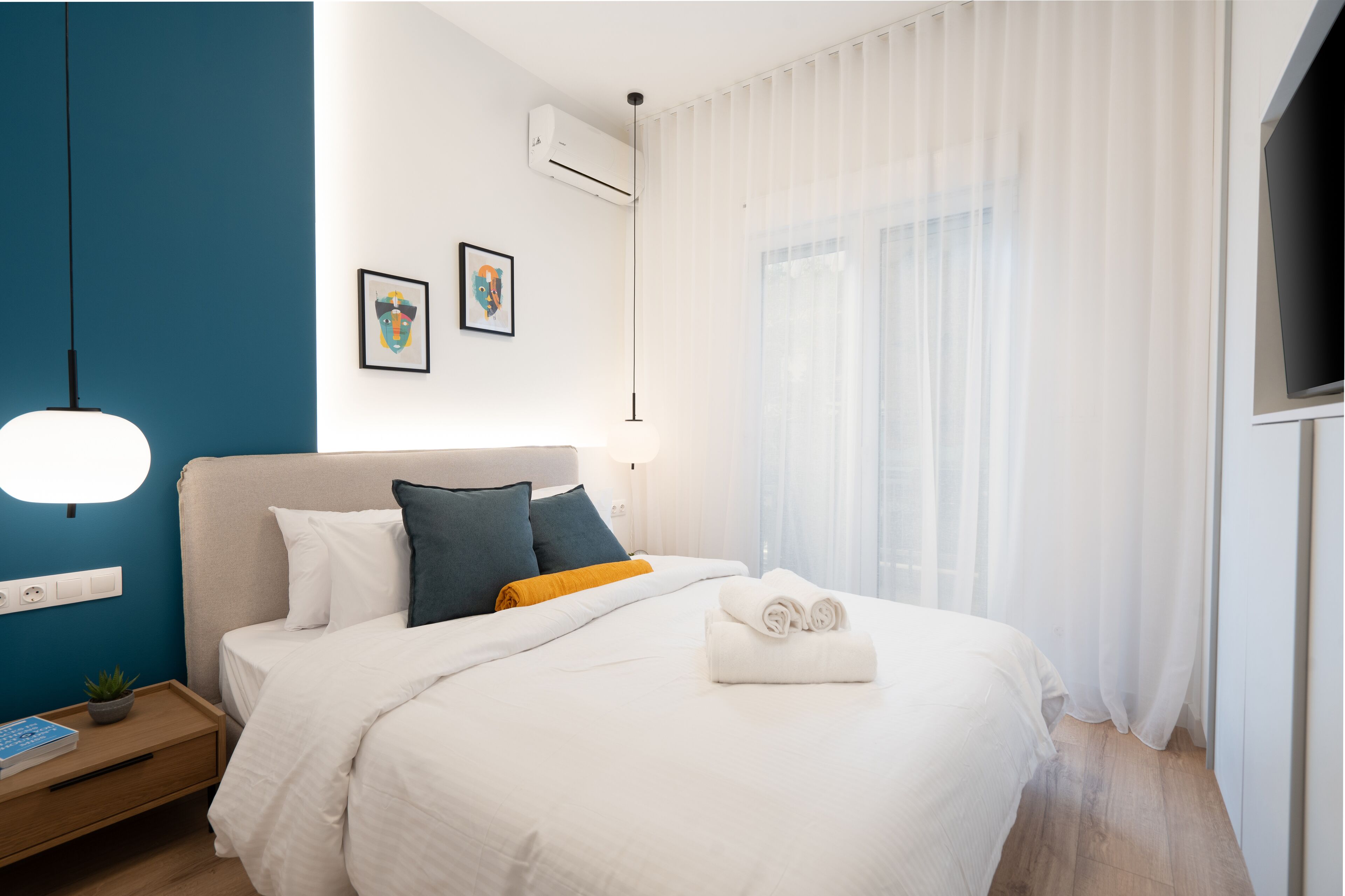 Ein modernes Hotelzimmer mit einem großen Bett, einem Nachttisch mit Lampe, einem Fernseher und einem Fenster mit durchsichtigen Vorhängen.
