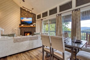 A cozy living room with a fireplace, a television mounted on a stone wall, and a dining area with a view of the outdoors.