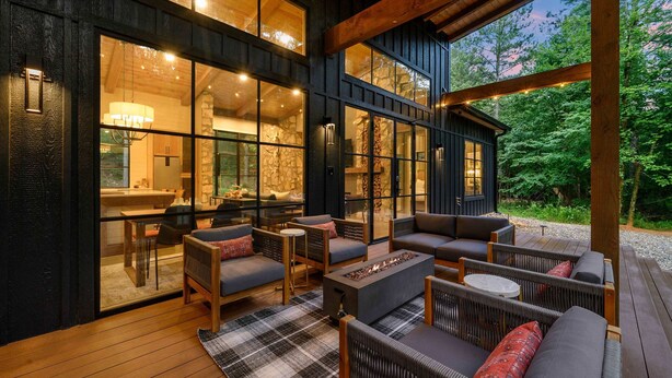Un espacio al aire libre acogedor con una chimenea, mobiliario de madera y grandes ventanales con vistas a un bosque.