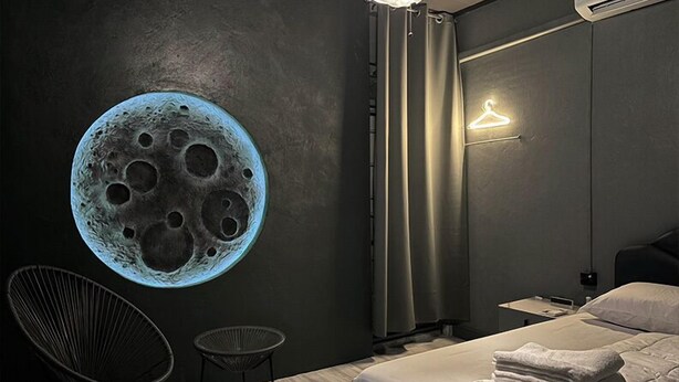A bedroom with a large wall mural of the moon, a bed with neatly folded towels, a chair, and a hanging light fixture. - Doctor Alien Hostel (Bangkok)